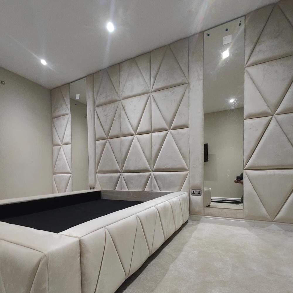 The image depicts a modern wall panel, minimalistic bedroom interior featuring geometric patterns. The room's focal point is a bed with a broad, cushioned frame covered in cream-colored upholstery with a triangular stitch pattern. The headboard wall matches the bed with large, upholstered panels forming a repeating pattern of triangles. A tall mirror is embedded in the wall to the right of the bed. 