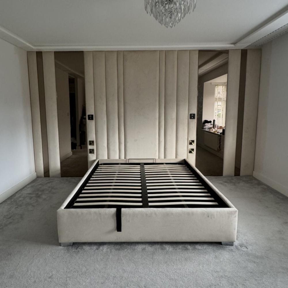 The image shows a modern, minimalist bedroom interior. The room features a large bed frame without a mattress, situated centrally on a plush, light-grey carpet. The wall panels bed frame is upholstered in a similar grey tone, adding a uniform appearance. Behind the bed, the wall is adorned with vertically aligned, padded beige panels, flanked by mirrored sections, contributing to a sense of depth and spaciousness. 