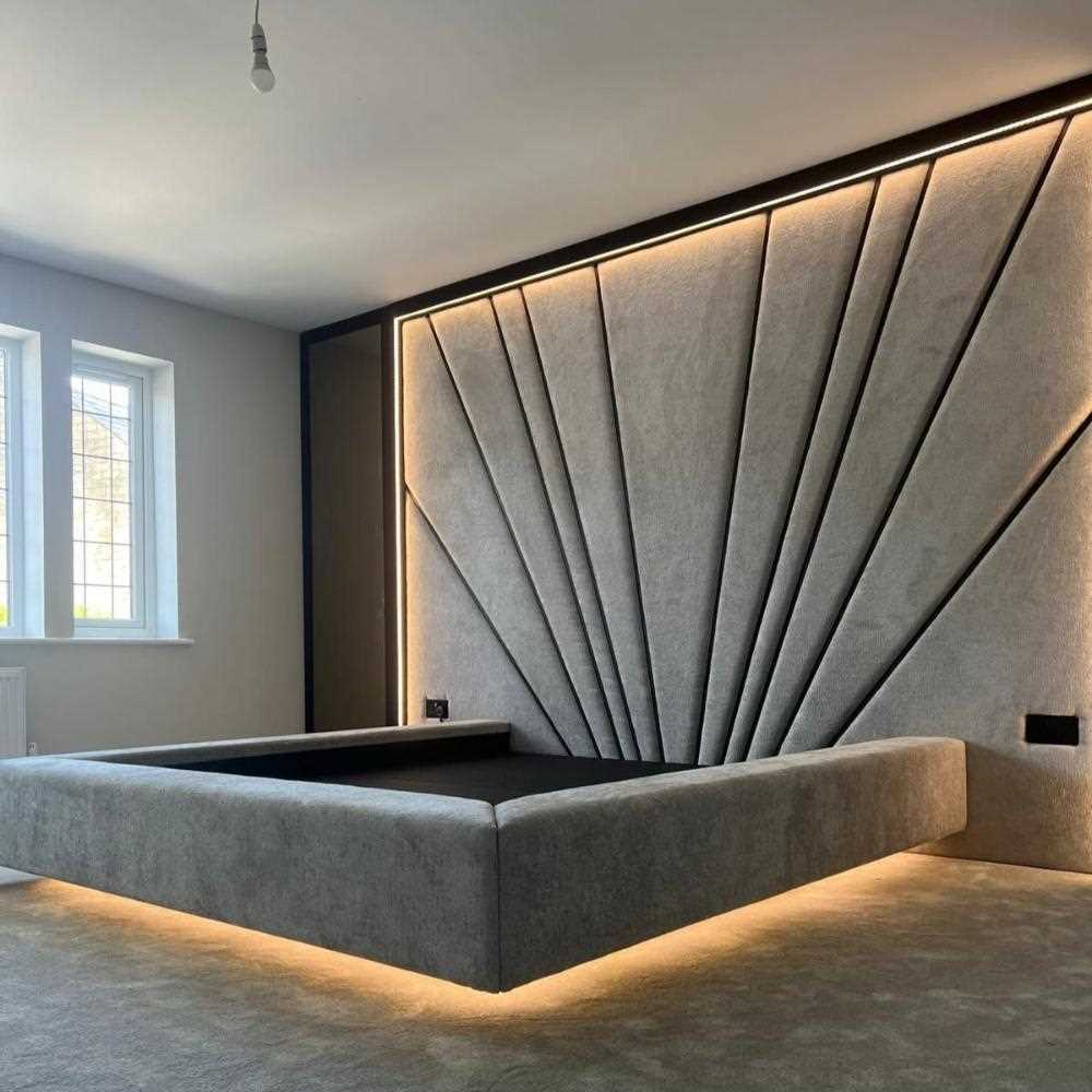 The image shows a modern, minimalist bedroom featuring a wall Panels bed with a striking architectural design. The bed frame appears to be upholstered in a soft gray fabric, suspended slightly above the floor, giving it a floating appearance. It is accentuated by a warm white underglow from lighting beneath the bed. The headboard is made of cushioned panels that form a fan-like geometric pattern, with each panel extending from a central point outward. 