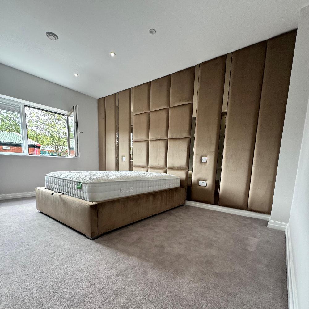 The image portrays a wall panels modern, minimalist bedroom with a focus on a large bed centered against an accent wall. The bed has a tall, plush, light brown padded headboard extending to the ceiling, providing a textured backdrop. The broad headboard is composed of large rectangular panels, creating a sophisticated pattern. A mattress rests on the bed frame, appearing firm and well-fitted. The room features light gray carpeting, complementing the neutral tones of the walls and furniture.