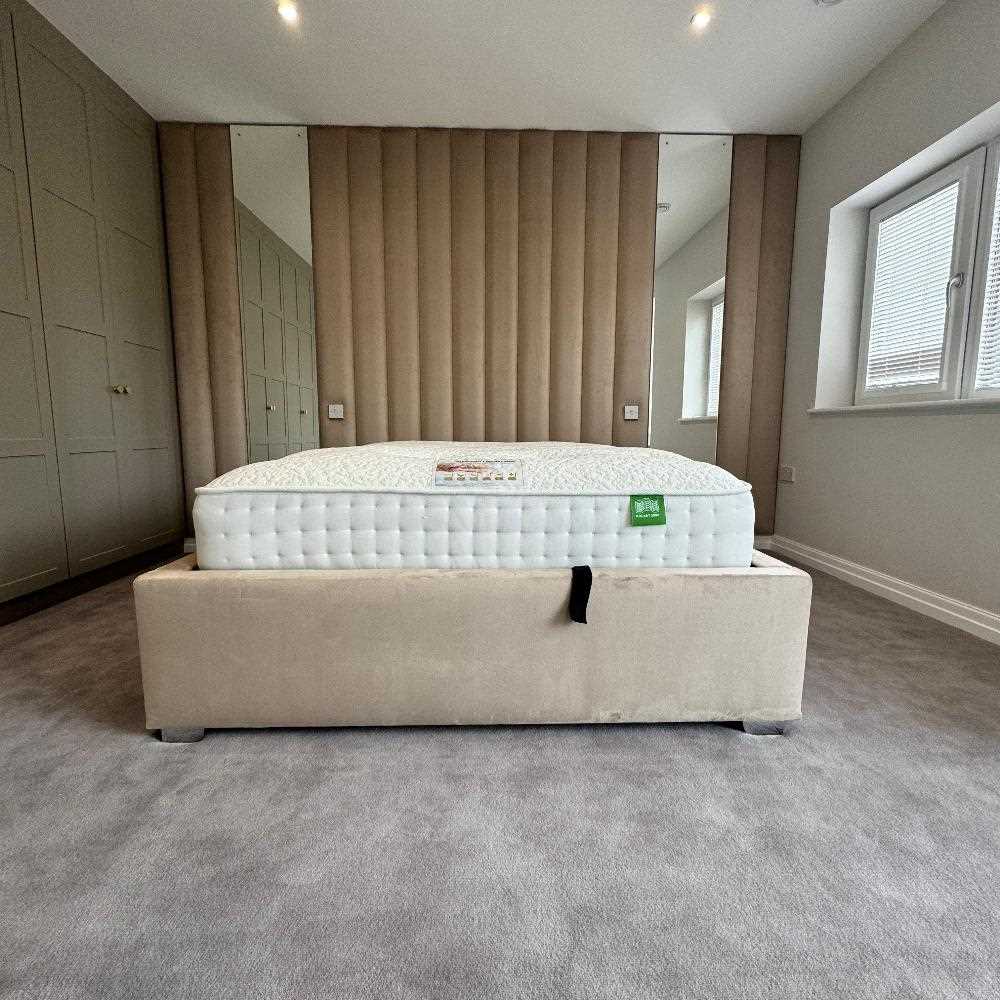 The image shows a bedroom with a plush, white mattress on a beige upholstered bed frame. The wall panels bed is positioned in front of a wall with a unique vertical panel design. The paneling is beige and adds a touch of texture and dimension to the room. The room is well-lit, with soft lighting coming from the ceiling. The floor is carpeted in a light grey color, creating a clean and minimalist aesthetic. The room also features two large mirrors, which reflect the surrounding space.