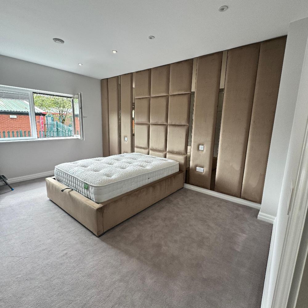 The image depicts a modern bedroom interior. The main focus is a large wall panels bed with a padded headboard against a wall. The bed frame and headboard are covered in a light brown suede-like material. The wall behind the bed features tall, vertical padded panels in the same brown material, giving a textured and luxurious appearance. On the floor is light grey carpet, adding to the room's soft aesthetic. 