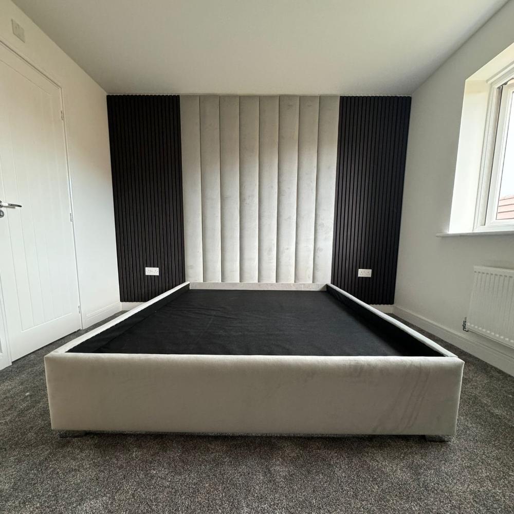 The image depicts a modern wall panels bedroom with a minimalist design. At the center is a large bed frame with a low profile and light gray upholstery. The headboard is composed of vertically aligned, padded panels in a soft gray shade, flanked by dark, textured panels on each side which create a striking contrast. The floor is carpeted in a muted gray tone. 