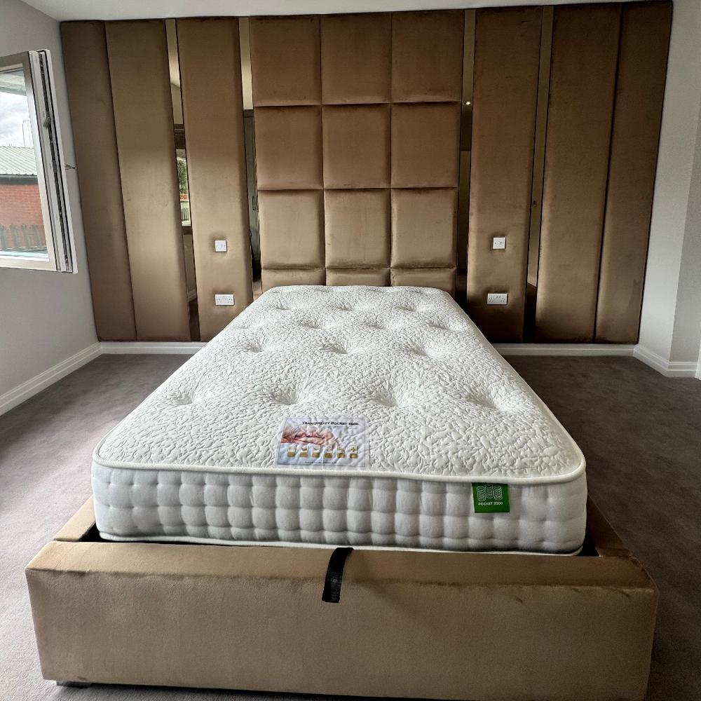 The image depicts a modern bedroom featuring a central mattress placed on a taupe-colored upholstered bed frame. The mattress has a textured white surface with a quilted pattern. A small, transparent wrapper with a red and white image is visible on the mattress. Above the bed, the wall is covered with large, padded, square panels in a bronze color, forming a grid pattern. The room has beige carpeting, and an open window is visible on the left, allowing natural light to illuminate the room.