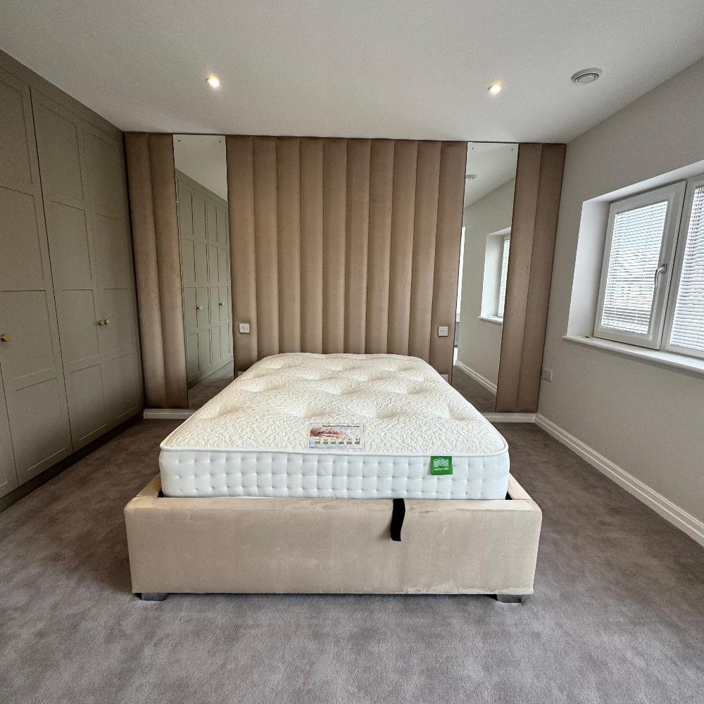 The image shows a modern bedroom with a beige upholstered bed frame. The bed is positioned in front of a wall with vertical wall panels, creating a visually appealing and textured backdrop. The mattress is white and features a green label with a logo on its top surface. The bedroom has a light grey carpet and a large window that lets in natural light. The walls are painted in a neutral color, and the overall design is clean and minimalist. 