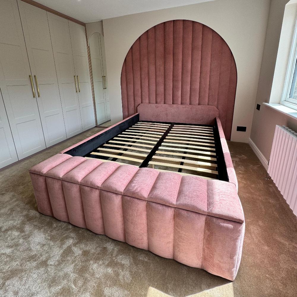 The image depicts a bedroom with a prominent wall panel bed frame in the center. The bed frame features a plush, padded, pink fabric with a ribbed texture. The headboard is large, extending upward in an arched shape, continuing the ribbed pattern. The bed's baseboard is similarly upholstered. The mattress is absent, revealing horizontal wooden slats. To the left of the bed, there is a wall of off-white, paneled wardrobes with gold handles, adding a touch of elegance. 