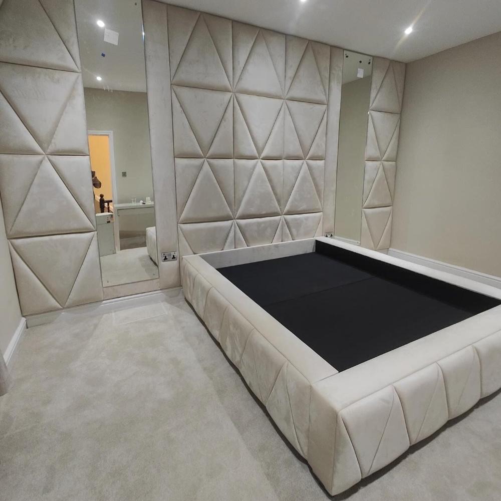 The image depicts a modern, minimalist bedroom with a distinctive design. The walls are covered in light beige, padded wall panels featuring a pattern of large, embossed triangular shapes. The flooring is covered in a matching light beige carpet. A large mirror is positioned on the left side of the room, reflecting part of the wall and an open doorway which reveals a different room with a person partially visible. 