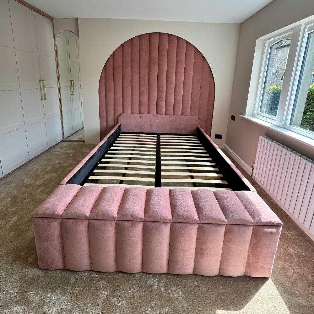 The image depicts a bedroom featuring a stylish, modern wall panels bed frame. The bed is upholstered in a soft pink fabric with vertical seams that add a textured pattern to the headboard and surrounding frame. The headboard is tall and semi-circular in shape, providing a bold visual focal point. The bed frame is currently without a mattress, exposing wooden slats that support the mattress. The room is carpeted in a light brown shade that complements the bed's color. 