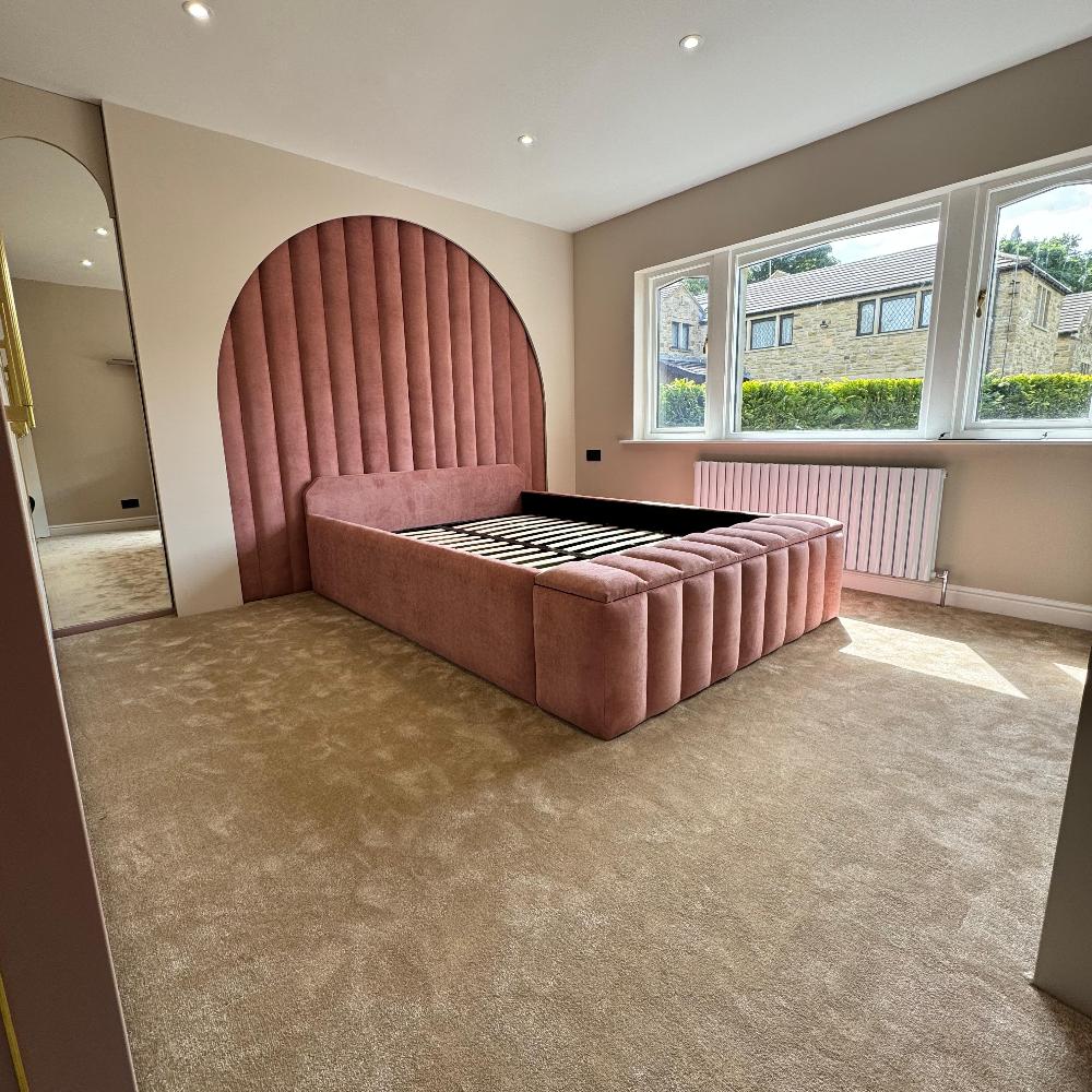 The image shows a bedroom featuring a modern wall panel bed with a distinctive design. The room has beige carpet flooring and is well-lit with natural light coming from a large window on the right. The bed has a pink upholstered frame with a headboard shaped in a large, semi-circular arch that extends to the floor. The upholstery is vertically tufted, creating a textured pattern. To the left, there's a tall, narrow mirror that reflects part of the room. The walls are painted in a light, neutral color. 