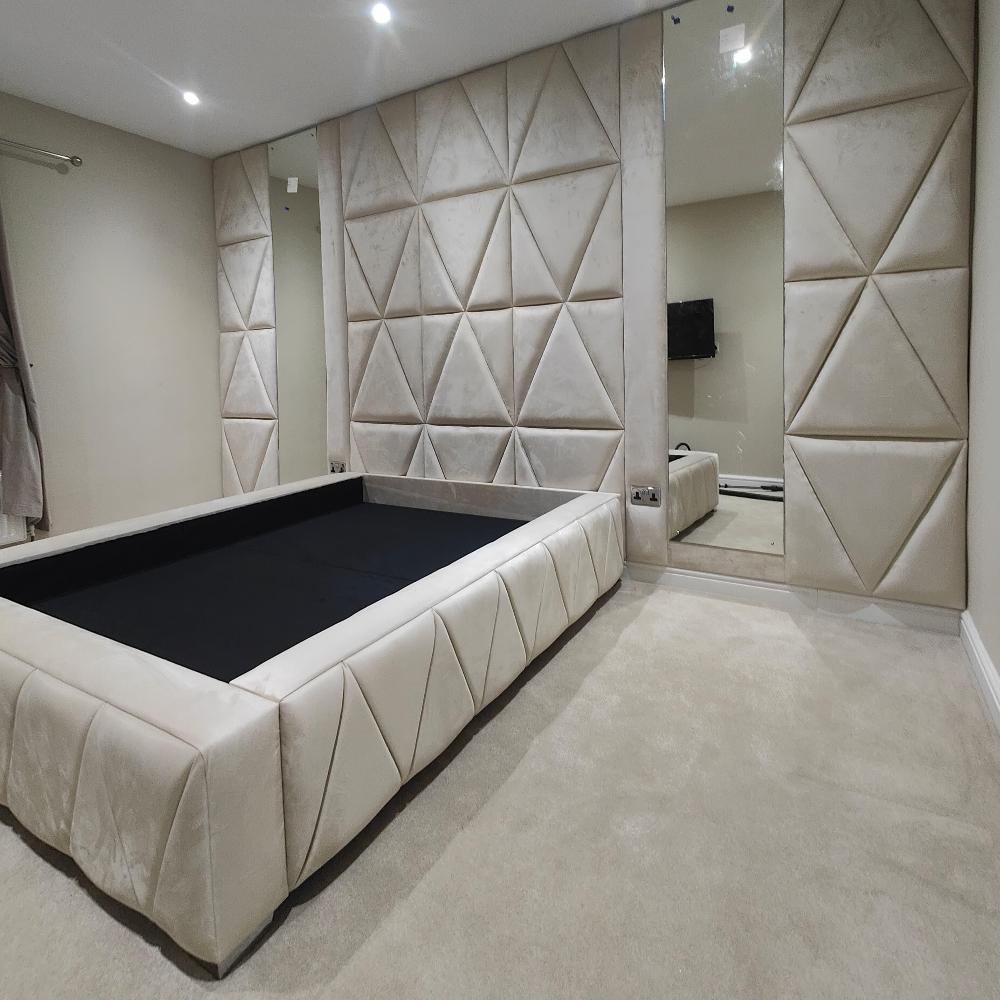 The image shows a modern wall panel minimalistic bedroom with beige and black tones. The dominant feature is a large, padded bed frame with a black mattress. The frame is upholstered in a soft, beige fabric with a subtle triangular pattern. The walls are adorned with large, mirrored panels reflecting room's features. The central wall behind the bed is covered with beige, plush triangular panels matching the bed’s design. The flooring is a light beige carpet that complements the room's overall color scheme. 