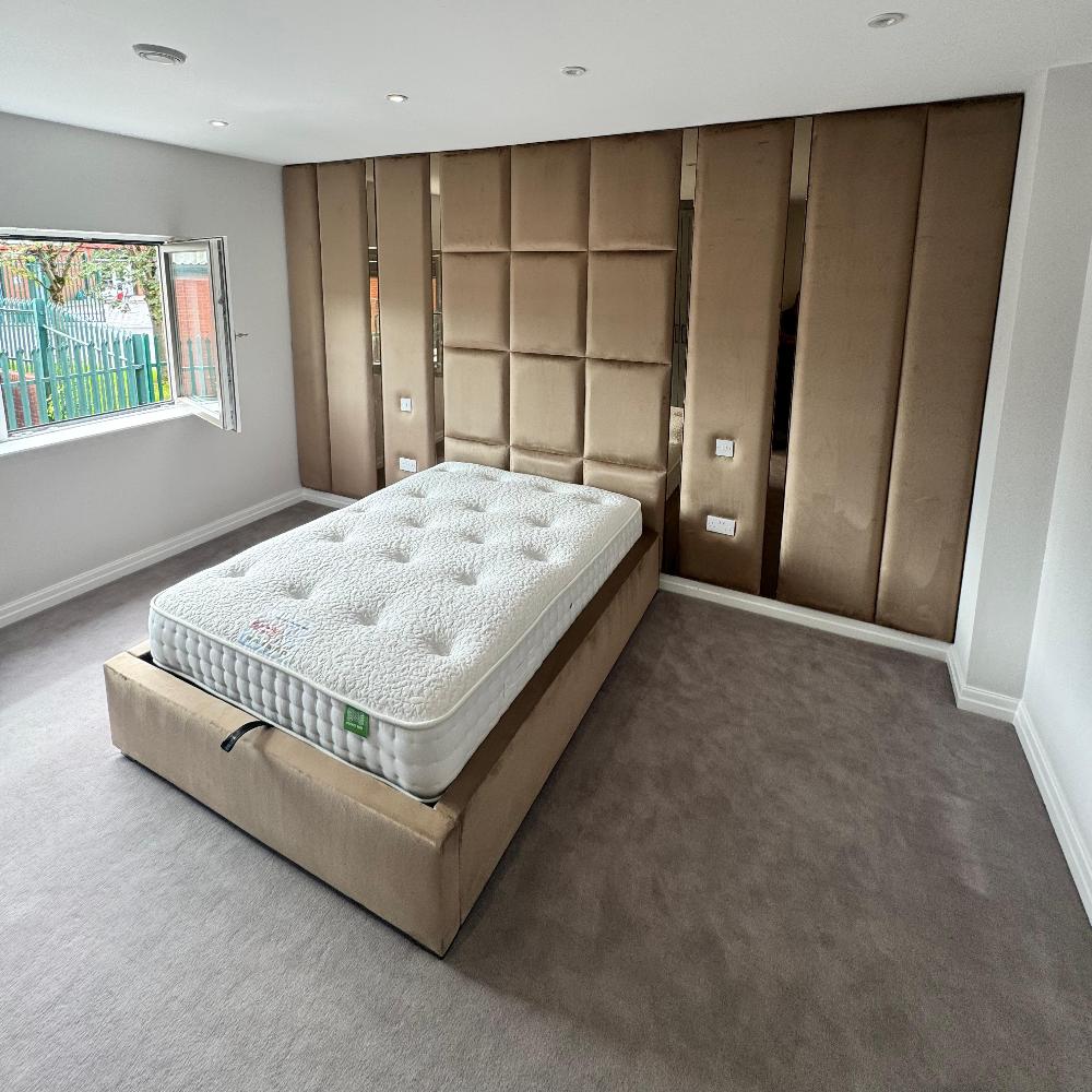 The image shows a modern bedroom with a focus on a large wall panels bed positioned centrally. The bed has a beige upholstered base with a white mattress on top, featuring a quilted pattern. The room has a plush grey carpet covering the floor. Behind the bed is a striking feature wall composed of vertically stacked, padded brown panels, adding texture and depth to the space. An open window to the left reveals a partial view of a garden and allows natural light to illuminate the room. 