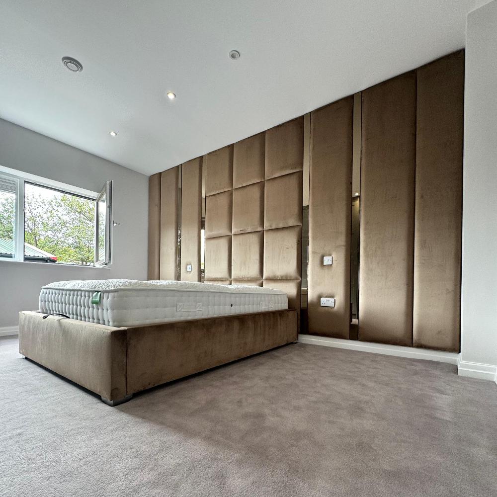 The image depicts a modern wall panel bedroom with a minimalist design. A large bed with a plush brown fabric frame and a white mattress occupies the foreground. The bed is positioned against an accent wall made of tall, vertical panels covered in brown fabric. The room is carpeted with a light gray rug, adding a soft texture to the floor. To the left, a window with white framing is open, allowing natural light to enter the room. The ceiling is white and contains small recessed lights.