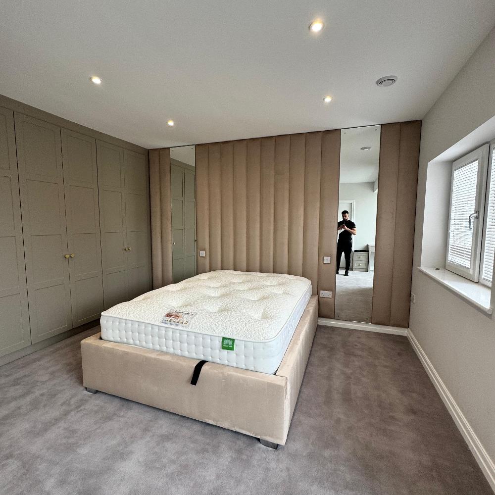 The image shows a modern bedroom with a beige tufted headboard and a plush white mattress. The wall panels bed is positioned against a wall that is covered in mirrored panels. The room is spacious and well-lit, with a large window providing natural light. There are multiple recessed lights in the ceiling, and the floor is covered in soft, gray carpet. The room is also decorated with a set of double doors with a light brown finish. The overall impression is one of luxury and comfort.  
