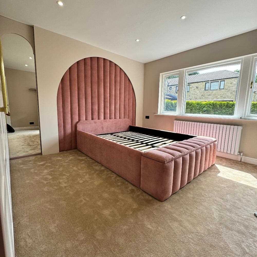 The image shows a bedroom featuring a large, upholstered bed frame without a mattress. The bed is positioned against a wall panels with a prominent, semi-circular, padded headboard that extends nearly to the ceiling. The upholstery is a soft, dusty rose color, with vertical stitching patterns visible on the headboard and footboard. The room has beige carpeting and light-colored walls. 