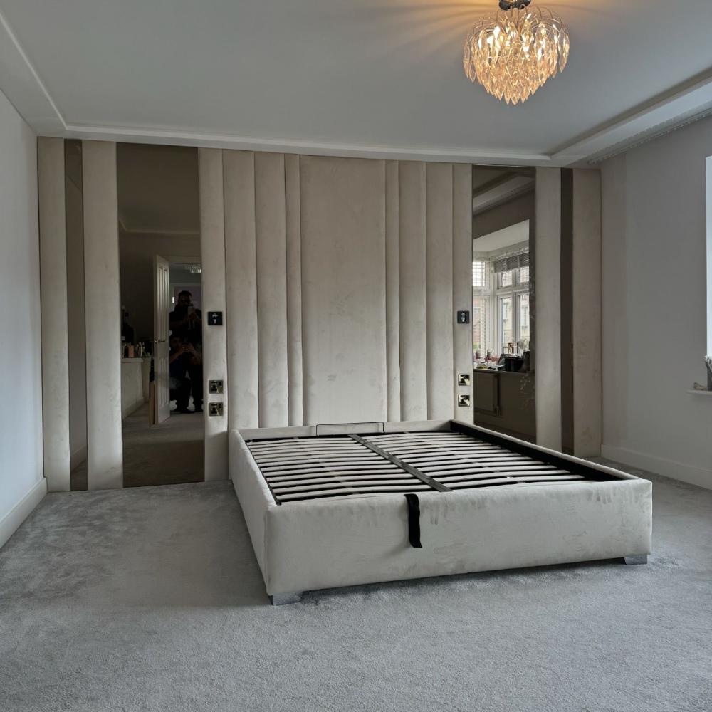 The image shows a modern, minimalist wall panels bedroom with a gray color scheme. Central to the room is a large, fabric-upholstered bed frame without a mattress, featuring horizontal slats. The headboard wall is composed of vertical panels in a soft beige hue, flanked by two tall, narrow mirrors that add depth to the space. Above the bed hangs a decorative chandelier with crystal-like elements, casting subtle light. The floor is covered in a matching gray carpet. 