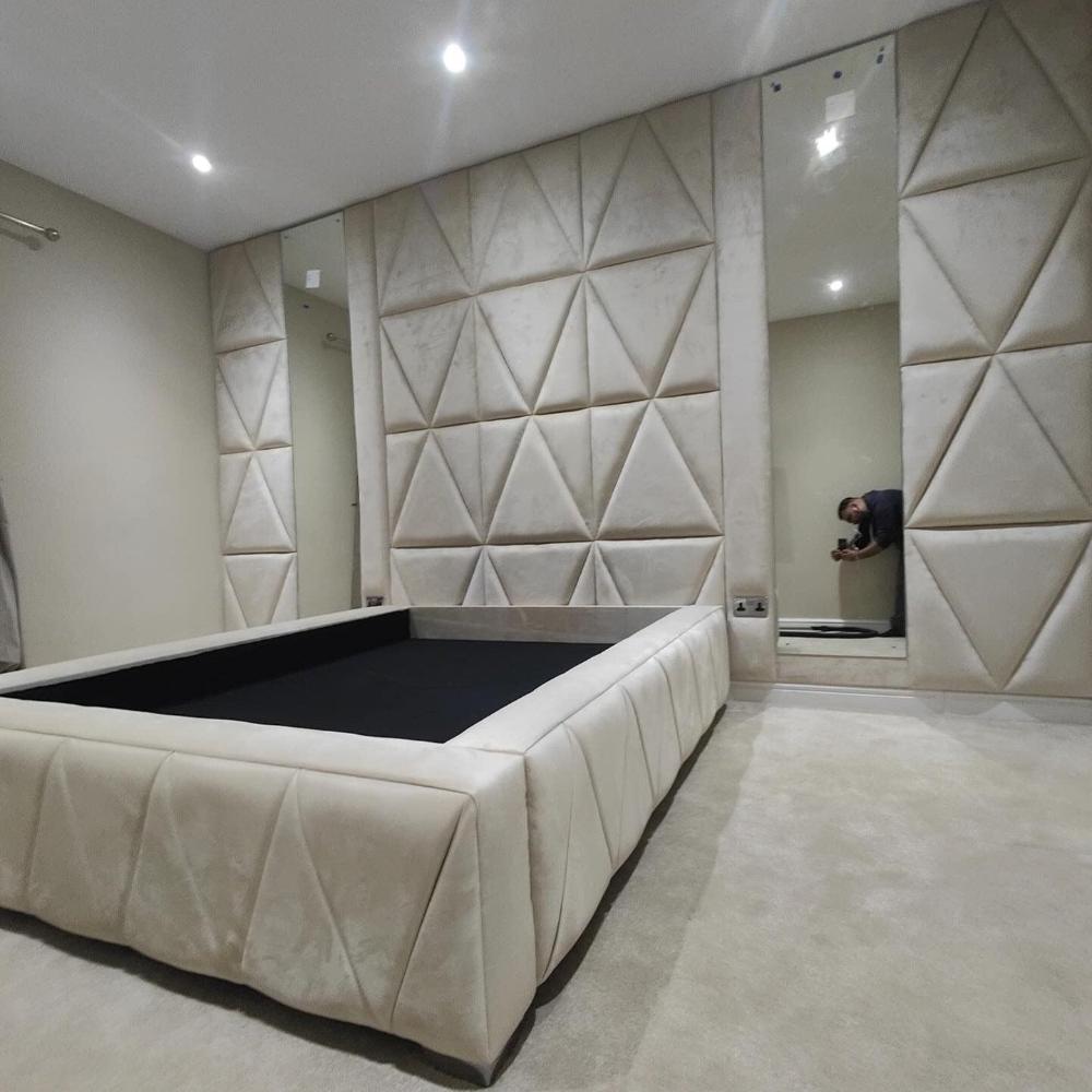 The image portrays a modern wall panel, minimalist bedroom with a strong geometric theme. The focal point is a large, rectangular bed frame with a soft, cream-colored upholstered surface that features subtle diagonal stitching. The walls are covered in expansive, padded panels designed with a pattern of large, soft triangles, complementing the texture of the bed. The room appears spacious with light-colored carpeting and muted beige walls, enhancing the serene ambiance.